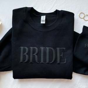 K&ouml;nnte beinhalten: Schwarzer Pullover mit dem Wort "BRIDE" in erhabener Stickerei. Ein zweiter schwarzer Pullover mit den Buchstaben "B" und "S" ist ebenfalls sichtbar, die mit einem Herz dazwischen gestickt sind.