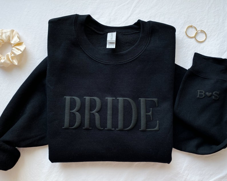 K&ouml;nnte beinhalten: Schwarzer Pullover mit dem Wort "BRIDE" in erhabener Stickerei. Ein zweiter schwarzer Pullover mit den Buchstaben "B" und "S" ist ebenfalls sichtbar, die mit einem Herz dazwischen gestickt sind.