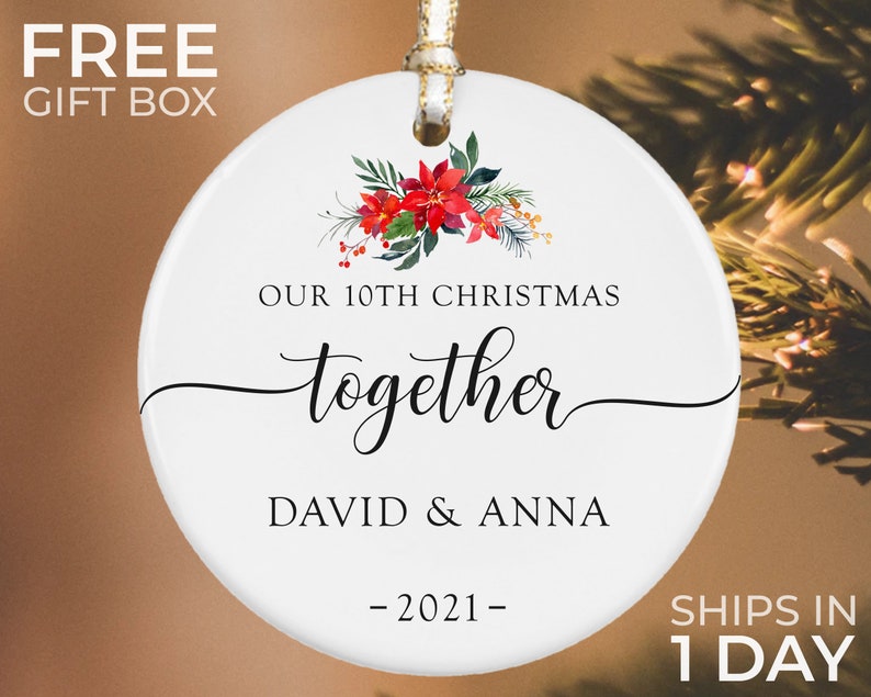 Puede incluir: Adorno navide&ntilde;o redondo de cer&aacute;mica blanca con lazo dorado.  Presenta texto "OUR 10TH CHRISTMAS together DAVID & ANNA -2021-" y motivos florales. Incluye una "FREE GIFT BOX".
