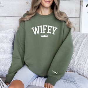 May include: A green sweatshirt with the text "WIFEY EST. 2024" in white. The sweatshirt has a white heart with an arrow on the left sleeve.