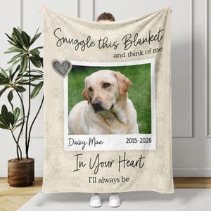 May include: A cream-colored fleece blanket featuring a central photo of a yellow Labrador retriever. The text reads Snuggle this Blanket and think of me, Daisy Mae, 2015-2026, and In Your Heart I'll always be. A gray heart and paw print patterns are visible.