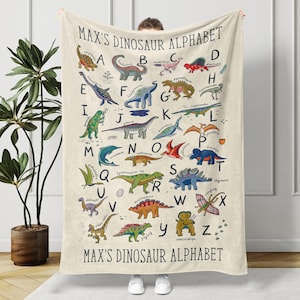 May include: A cream-colored blanket featuring a colorful dinosaur alphabet. Each letter of the alphabet is paired with a different dinosaur illustration. The text "MAX'S DINOSAUR ALPHABET" is printed at the top and bottom of the blanket.
