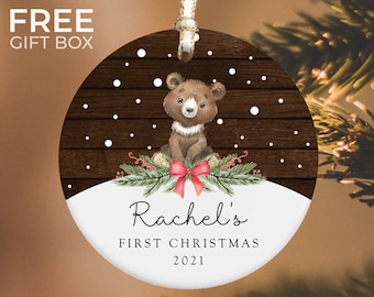 May include: A round, white Christmas ornament with a brown wooden background and a cute cartoon bear wearing a red bow. The ornament says "Rachel's First Christmas 2021".