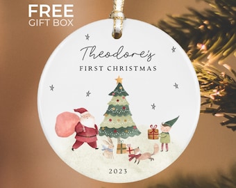 May include: White ceramic Christmas ornament with a watercolor illustration of Santa Claus, a Christmas tree, an elf, a bunny, and a fox. The ornament says "Theodore's First Christmas" and "2023".