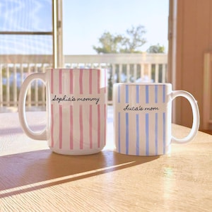 May include: Two ceramic mugs with handles. The larger mug features vertical pink stripes and the text "Sophia's mommy" in black script. The smaller mug features vertical blue stripes and the text "Luca's mom" in black script. Both mugs are white.