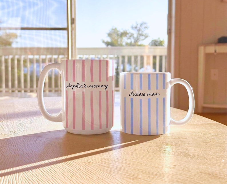 May include: Two ceramic mugs with handles. The larger mug features vertical pink stripes and the text "Sophia's mommy" in black script. The smaller mug features vertical blue stripes and the text "Luca's mom" in black script. Both mugs are white.