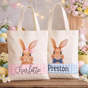 May include: Two cream-colored tote bags with bunny illustrations. One bunny has a pink bow, the other a blue bow tie. The first bag says "Charlotte" on a pink gingham stripe, the second says "Preston" on a blue gingham stripe. Easter-themed.