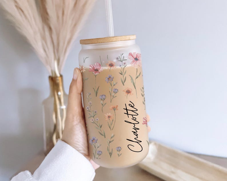 K&ouml;nnte beinhalten: Mattierter Glasbecher mit floralem Design mit rosa, lila und gelben Wildblumen. Der Becher enth&auml;lt einen Bambusdeckel und einen Strohhalm. Der Name "Charlotte" ist in einer Schreibschrift auf der Seite geschrieben.
