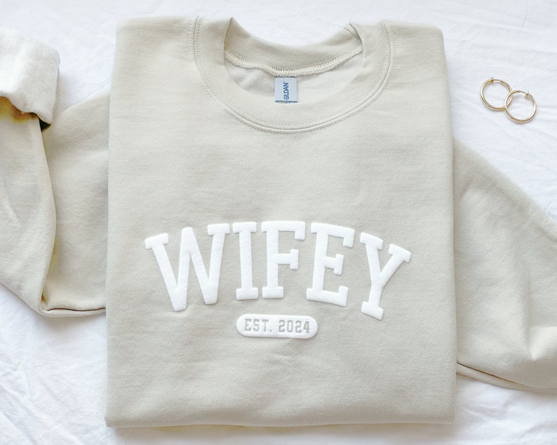May include: A light beige sweatshirt with the word "WIFEY" in white block letters and the text "EST. 2024" below it.