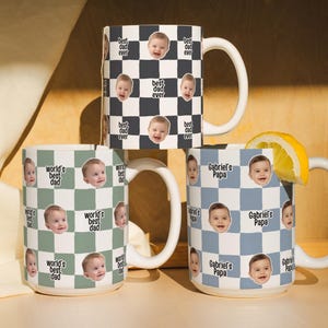 May include: Three white ceramic mugs with a checkered pattern and baby faces. One mug says "best dad ever", another says "world's best dad", and the third says "Gabriel's Papa". A lemon slice rests on one mug.