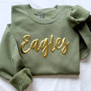 May include: An olive green sweatshirt with the word "Eagles" in gold script across the front. The sleeves are cuffed, and the crew neck sweatshirt is folded neatly on a white surface.