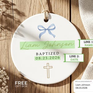 Peut inclure: Ornement de bapt&ecirc;me en c&eacute;ramique blanche avec un n&oelig;ud bleu, le nom "Liam Johnson" et la date "08.23.2026". Une croix dor&eacute;e est en bas. Le texte "BAPTIS&Eacute;" est &eacute;galement pr&eacute;sent.