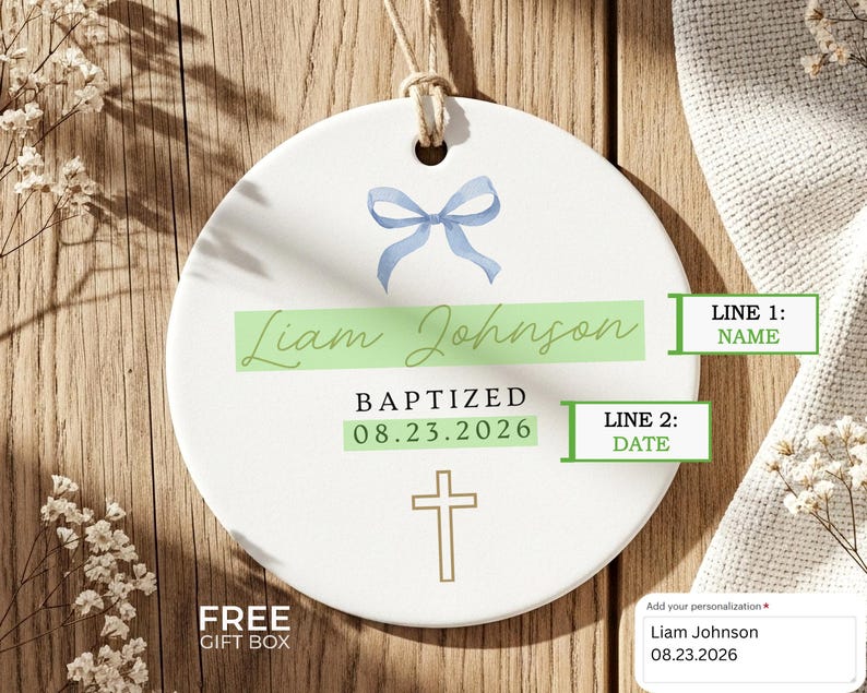 Peut inclure: Ornement de bapt&ecirc;me en c&eacute;ramique blanche avec un n&oelig;ud bleu, le nom "Liam Johnson" et la date "08.23.2026". Une croix dor&eacute;e est en bas. Le texte "BAPTIS&Eacute;" est &eacute;galement pr&eacute;sent.