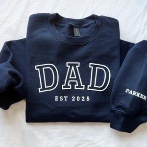 May include: A navy blue sweatshirt with the word "DAD" in white block letters and the text "EST 2025" below. The sweatshirt has a long sleeve and a crew neck.