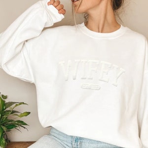 May include: White sweatshirt with the word "WIFEY" in raised lettering.