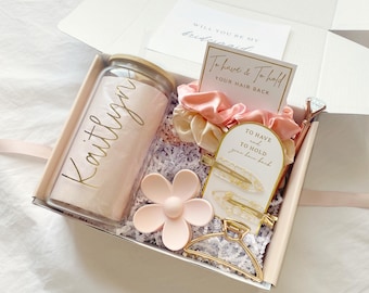 May include: A gift box containing a glass tumbler with a bamboo lid and the name Kaitlyn in gold script, two satin scrunchies, a matte pink flower hair clip, a gold metal hair claw, two gold-flecked hair clips, and a diamond-topped pen.