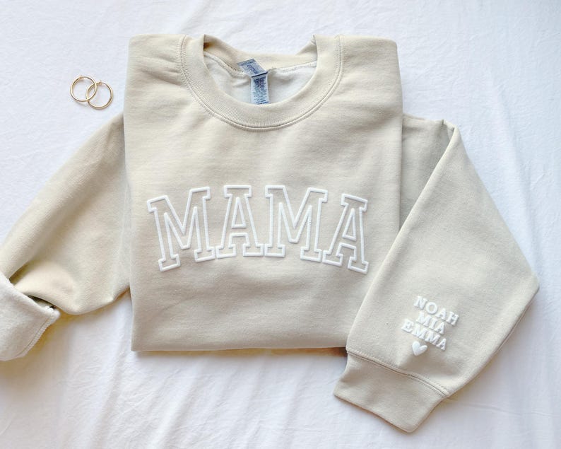 A sand colored sweatshirt with the word 'MAMA' in white outlined letters on the front. The right sleeve has kids names in white letters with a small white heart below the names.