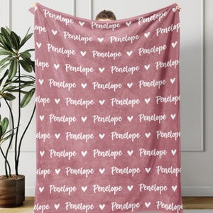 May include: A dusty rose blanket with the name "Penelope" repeated in a white script font, interspersed with white hearts. The blanket is soft and cozy, perfect for snuggling.