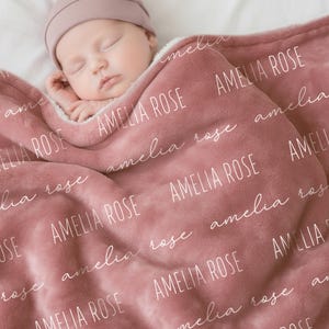 Może przedstawiać: Miękki kocyk dziecięcy w kolorze przygaszonego r&oacute;żu z powtarzającym się białym napisem AMELIA ROSE, wykonanym czcionką bezszeryfową oraz kursywą. Pluszowa tkanina ma wyraźną fakturę i jest narzucona na śpiące niemowlę.