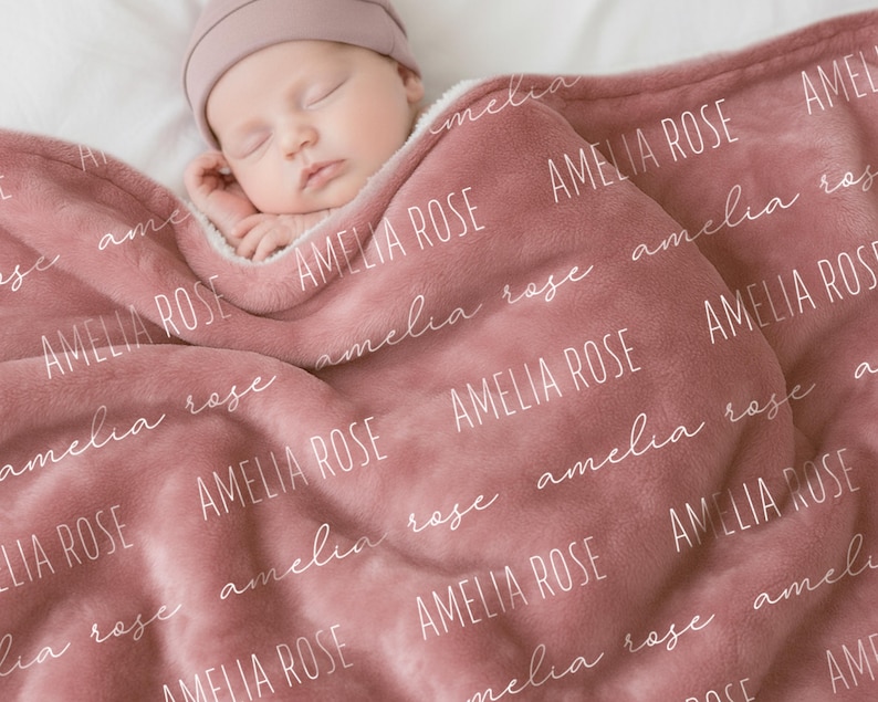 Może przedstawiać: Miękki kocyk dziecięcy w kolorze przygaszonego r&oacute;żu z powtarzającym się białym napisem AMELIA ROSE, wykonanym czcionką bezszeryfową oraz kursywą. Pluszowa tkanina ma wyraźną fakturę i jest narzucona na śpiące niemowlę.
