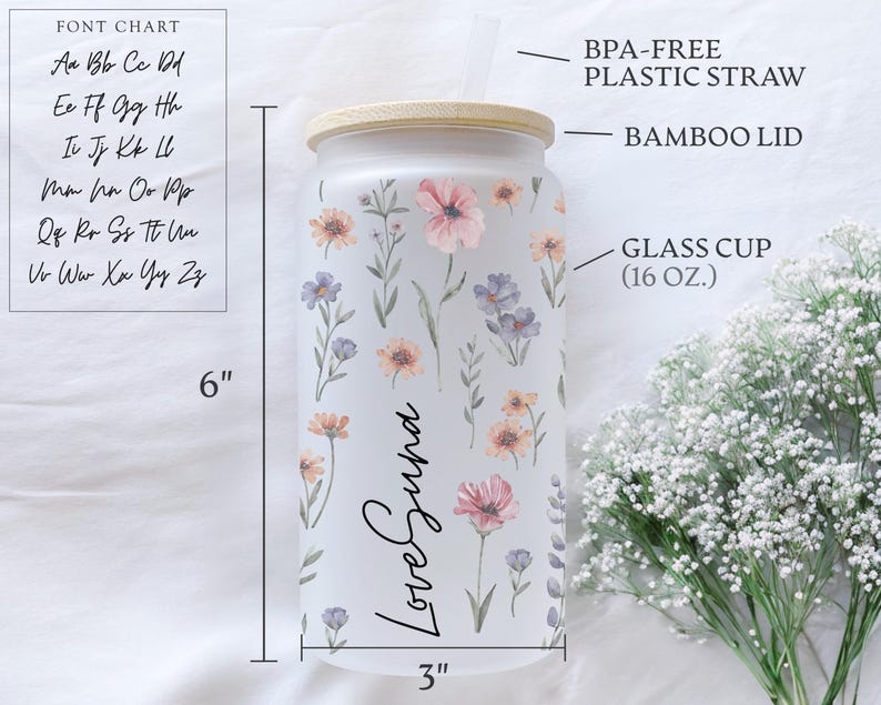 K&ouml;nnte beinhalten: Ein gefrosteter Glasbecher mit Bambusdeckel, verziert mit pastellfarbenen Blumen. Der Becher ist 15 cm hoch und 7,6 cm breit, mit dem Text "Love Suna". Enth&auml;lt einen BPA-freien Kunststoffstrohhalm.