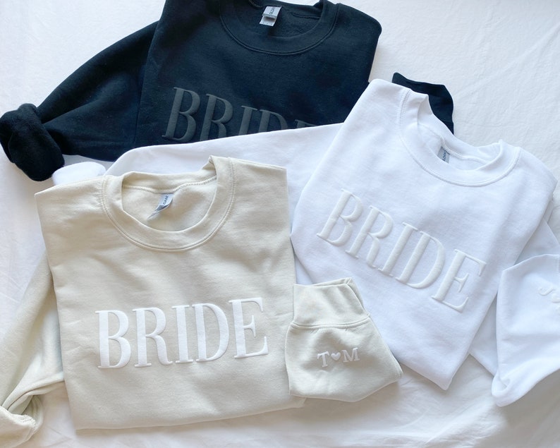 K&ouml;nnte beinhalten: Drei Sweatshirts mit dem Wort "BRIDE" bestickt. Die Sweatshirts sind in Schwarz, Wei&szlig; und Beige.