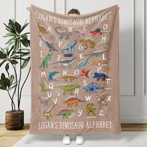 Könnte beinhalten: Eine beige Decke mit einem Dinosaurier-Alphabet-Design. Die Decke zeigt farbenfrohe Illustrationen verschiedener Dinosaurier, jeweils gepaart mit einem Buchstaben des Alphabets. Der Text "LOGAN'S DINOSAUR ALPHABET" ist oben und unten aufgedruckt.