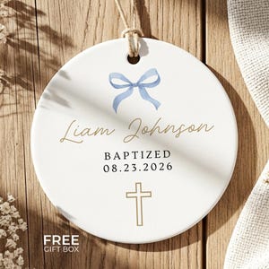 Peut inclure: Un souvenir de bapt&ecirc;me rond en c&eacute;ramique blanche. Il pr&eacute;sente un n&oelig;ud bleu, le nom "Liam Johnson" en &eacute;criture dor&eacute;e et le texte "BAPTIS&Eacute; 08.23.2026". Une croix dor&eacute;e est &eacute;galement pr&eacute;sente. Une ficelle est attach&eacute;e pour l'accrocher.