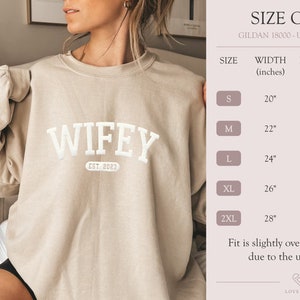 May include: Beige sweatshirt with the text "WIFEY EST. 2023" in white. Size chart for Gildan 18000 unisex sweater with measurements in inches.
