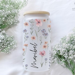 K&ouml;nnte beinhalten: Mattierter Glasbecher mit Bambusdeckel, verziert mit Aquarell-Blumendesigns in Rosa, Orange und Lila. Der Name "Maribel" ist in eleganter Schrift geschrieben. Enth&auml;lt einen Strohhalm.