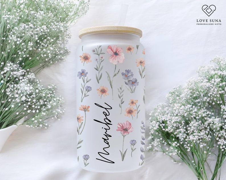 K&ouml;nnte beinhalten: Mattierter Glasbecher mit Bambusdeckel, verziert mit Aquarell-Blumendesigns in Rosa, Orange und Lila. Der Name "Maribel" ist in eleganter Schrift geschrieben. Enth&auml;lt einen Strohhalm.