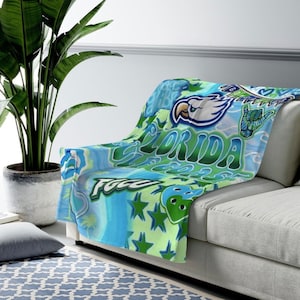 May include: A blue and green fleece throw blanket with a Florida theme. The blanket features a white bird, a green palm tree, and the words "Florida Gulf Coast".