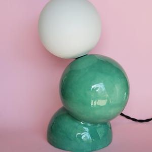 Puede incluir: Una lámpara de cerámica moderna con una esfera blanca sobre una esfera verde azulado brillante, apilada sobre una base verde azulado más pequeña. La lámpara tiene un cable de alimentación negro y está sobre un fondo rosa.