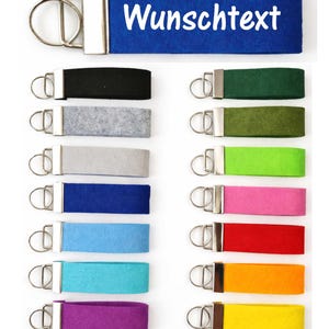 Könnte beinhalten: Eine Auswahl an farbigen Schlüsselanhängern mit Metallbeschlägen. Der obere Schlüsselanhänger ist blau mit dem weißen Text "Wunschtext". Weitere Schlüsselanhänger sind schwarz, grau, grün, oliv, limette, pink, rot, orange und gelb.