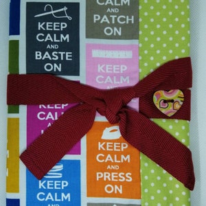 May include: A colorful fabric book with a red ribbon tied around it. The book has a patchwork design with different colored squares featuring the text "Keep Calm and..." followed by a sewing-related word, such as "Trim On", "Patch On", and "Press On".