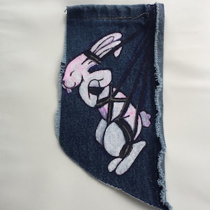 May include: A pink and white cartoon bunny is painted on a blue denim patch. The bunny is tied up with black ropes and has a pink blush on its cheeks.