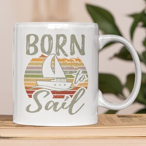 Peut inclure: Une tasse en céramique blanche avec un design de coucher de soleil rétro et le texte "Born to Sail" dans une police vintage. Une silhouette de voilier est représentée au centre du coucher de soleil.