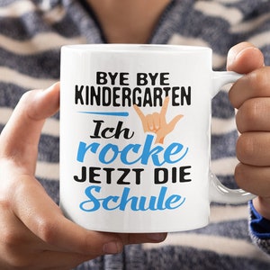 Puede incluir: Taza de cerámica blanca con un diseño azul y negro que dice "Bye Bye Kindergarten I rocke jetzt die Schule". La taza está siendo sostenida por la mano de una persona.