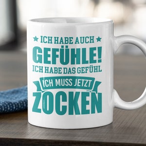 May include: White ceramic mug with a large handle, featuring teal text that reads "ICH HABE AUCH GEFÜHLE! ICH HABE DAS GEFÜHL ICH MUSS JETZT ZOCKEN". The mug also has a dishwasher safe symbol.