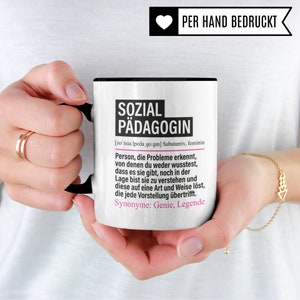 May include: White ceramic mug with a black handle. The mug has a black rim and a black line around the top. The mug has a black and white design with the text "SOZIAL P&Auml;DAGOGIN" and a definition of the word in German. The mug also has the text "PER HAND BEDRUCKT" in a heart shape.