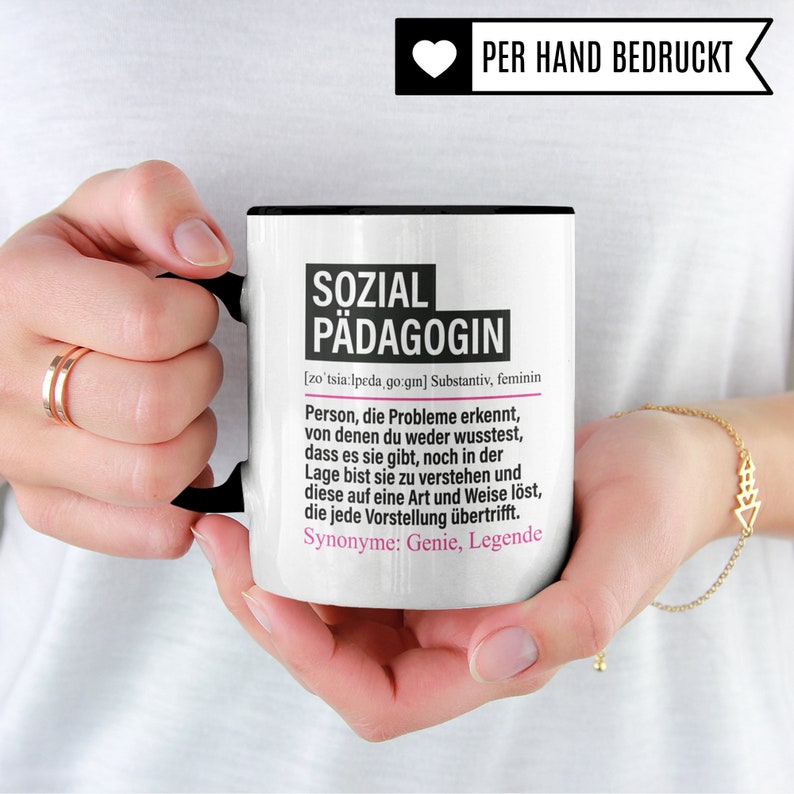 May include: White ceramic mug with a black handle. The mug has a black rim and a black line around the top. The mug has a black and white design with the text "SOZIAL P&Auml;DAGOGIN" and a definition of the word in German. The mug also has the text "PER HAND BEDRUCKT" in a heart shape.