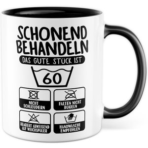 May include: White ceramic mug with a black rim and handle. The mug has black text that reads "SCHONEND BEHANDELN DAS GUTE STÜCK IST 60" and includes a washing instruction symbol with the number 60 inside. Below the symbol are four additional washing instruction symbols with text: "NICHT SCHLEUDERN", "FALTEN NICHT BÜGELN", "REAGIERT ABWEISEND AUF WEICHSPÜLER", and "HANDWÄSCHE EMPFOHLEN".