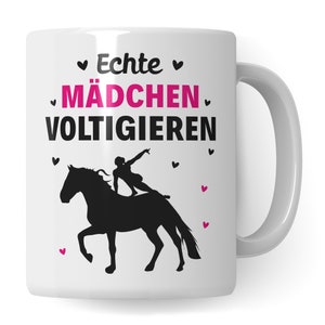 Op de afbeelding: Witte keramische mok met een zwart silhouet van een persoon die een voltige-oefening op een paard doet. De tekst "Echte MÄDCHEN VOLTIGIEREN" is in zwart met roze hartjes gedrukt.