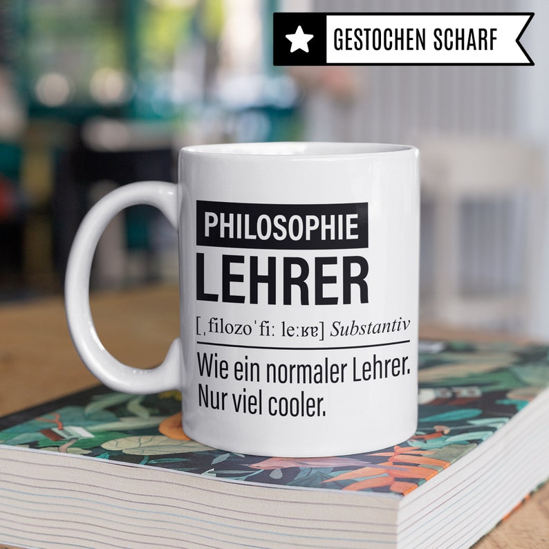 May include: White ceramic mug with a black design that reads "PHILOSOPHIE LEHRER [filozo'fi: le:ve] Substantiv Wie ein normaler Lehrer. Nur viel cooler."
