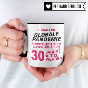 May include: White ceramic mug with a black rim and handle. The mug has a pink and black design with the text "SOGAR EINE GLOBALE PANDEMIE KONNTE MICH NICHT DAVON ABHALTEN 30 JAHRE ALT ZU WERDEN".