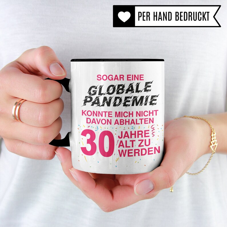 May include: White ceramic mug with a black rim and handle. The mug has a pink and black design with the text "SOGAR EINE GLOBALE PANDEMIE KONNTE MICH NICHT DAVON ABHALTEN 30 JAHRE ALT ZU WERDEN".