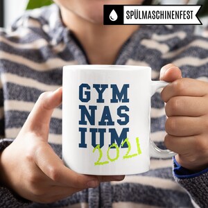 Pu&ograve; includere: Tazza in ceramica bianca con un design blu e verde. La tazza dice "GYMNASIUM 2021" in un carattere in grassetto.