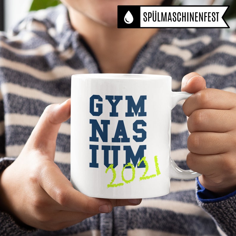 Pu&ograve; includere: Tazza in ceramica bianca con un design blu e verde. La tazza dice "GYMNASIUM 2021" in un carattere in grassetto.
