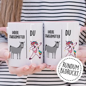 Peut inclure: Deux tasses en c&eacute;ramique blanche avec des bords roses. Les tasses pr&eacute;sentent une illustration en noir et blanc d'un cheval et d'une licorne de chaque c&ocirc;t&eacute;. Le texte sur les tasses est "Andere Schwiegerm&uuml;tter" et "Du". Les tasses sont tenues par les mains d'une personne. Un cercle rose avec le texte "RUNDUM BEDRUCKT" se trouve dans le coin inf&eacute;rieur droit de l'image.
