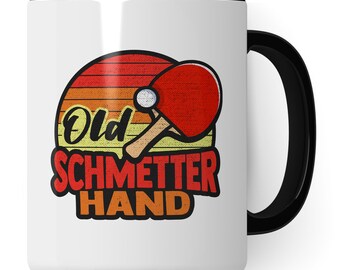 May include: White ceramic mug with a black handle. The mug features a retro-style design with a sunset background and a table tennis paddle and ball. The text "Old Schmetter Hand" is printed in yellow, orange, and red.